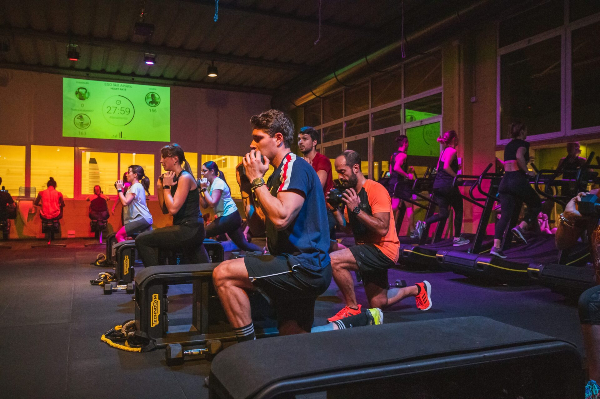 Gruppo di persone in allenamento funzionale, eseguendo affondi e sollevamento pesi in palestra.