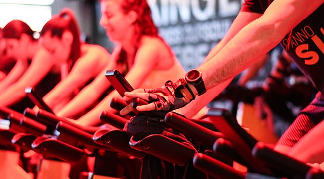 Gruppo di persone che partecipano a una lezione di spinning in palestra, mentre pedalano su biciclette stazionarie.