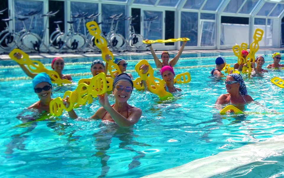 Gruppo di persone in piscina che partecipa a una lezione di acqua fitness, utilizzando attrezzi galleggianti gialli.