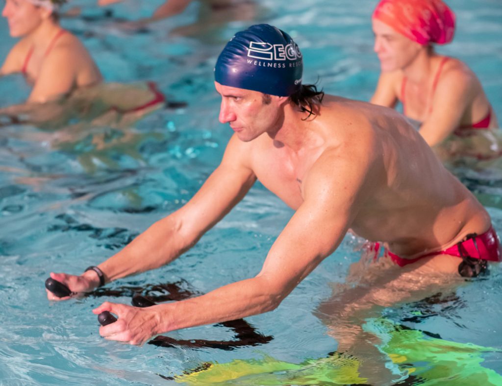 Uomo muscoloso in piscina, mentre esegue esercizi di acquagym utilizzando attrezzi galleggianti.