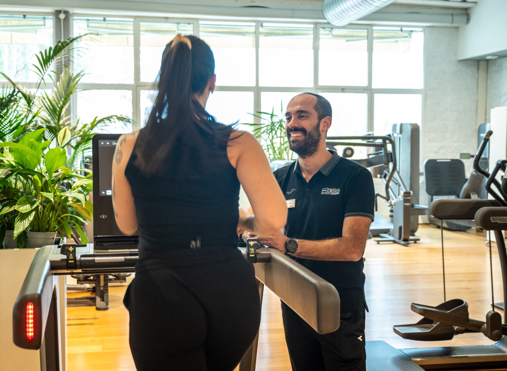 Istruttore in palestra che sorride mentre assiste una cliente su un tapis roulant.