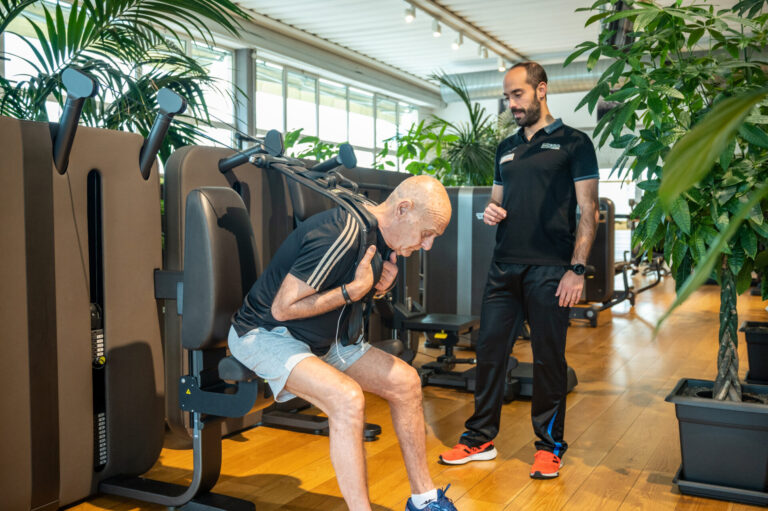 Un uomo anziano si allena su una macchina per il potenziamento muscolare sotto la supervisione di un istruttore in una palestra moderna.