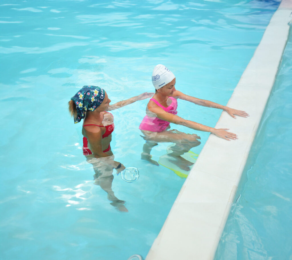 Istruttrice guida esercizi indrokinesi in piscina con una partecipante.