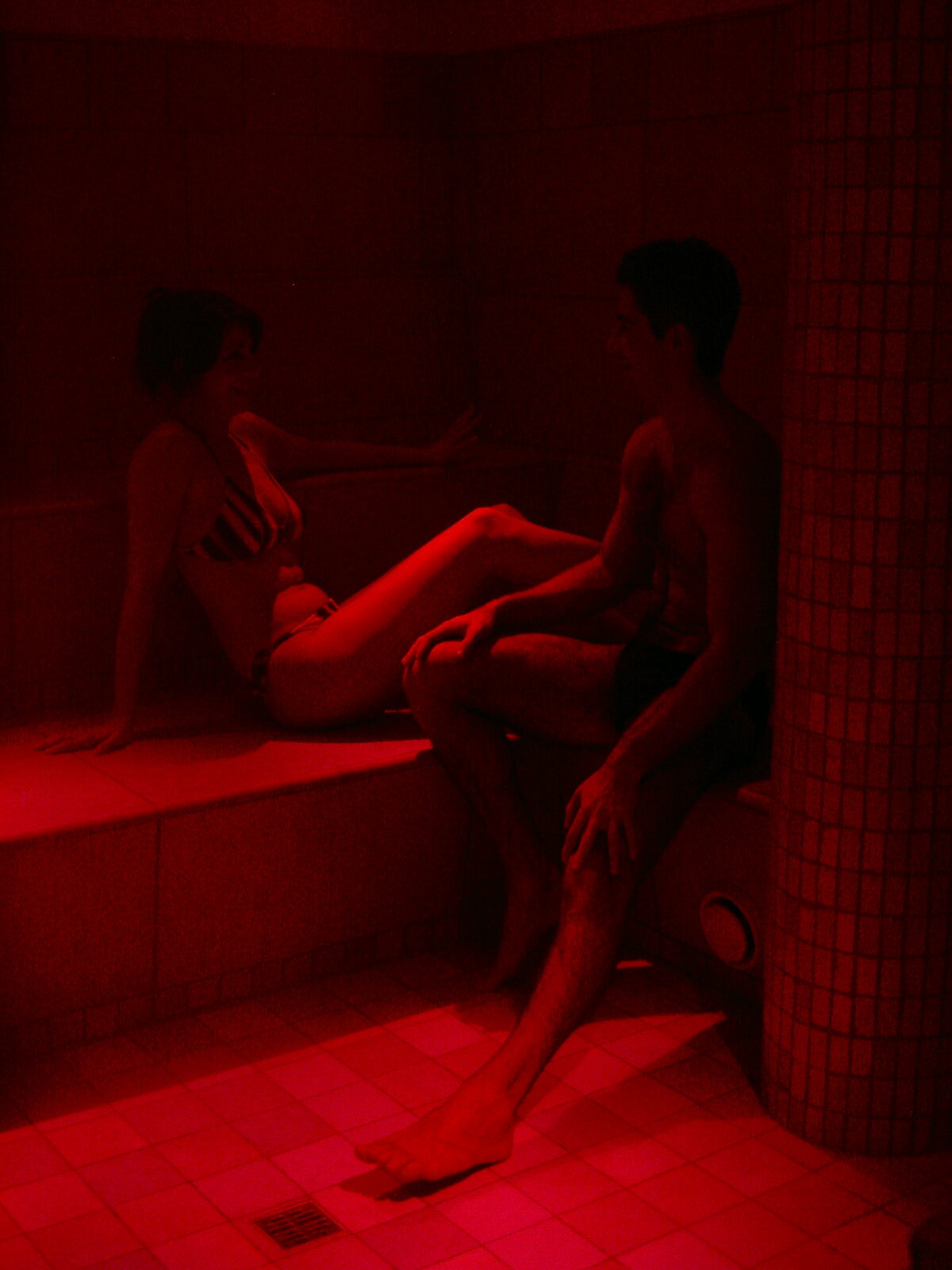 Due persone che si rilassano in un bagno turco illuminato da luce rossa, sedute su panche in un ambiente caldo.