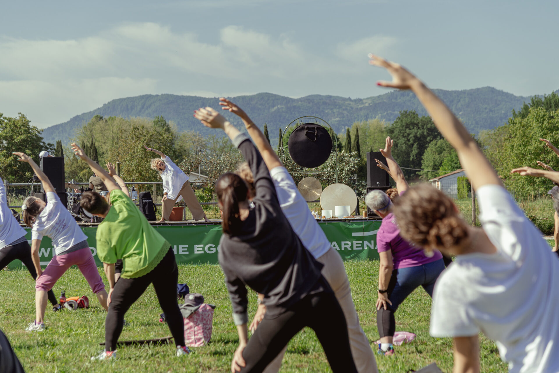 Partecipanti che eseguono esercizi di stretching guidati da un istruttore su un palco all'aperto.